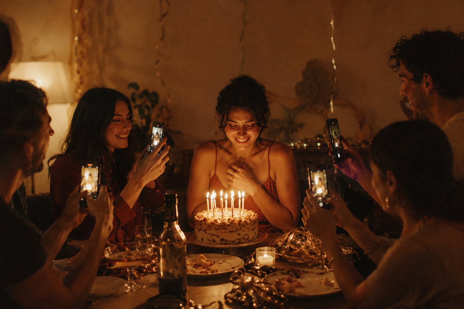 Friends filming a birthday cake with lit candles on their phones in a warm, dimly lit room