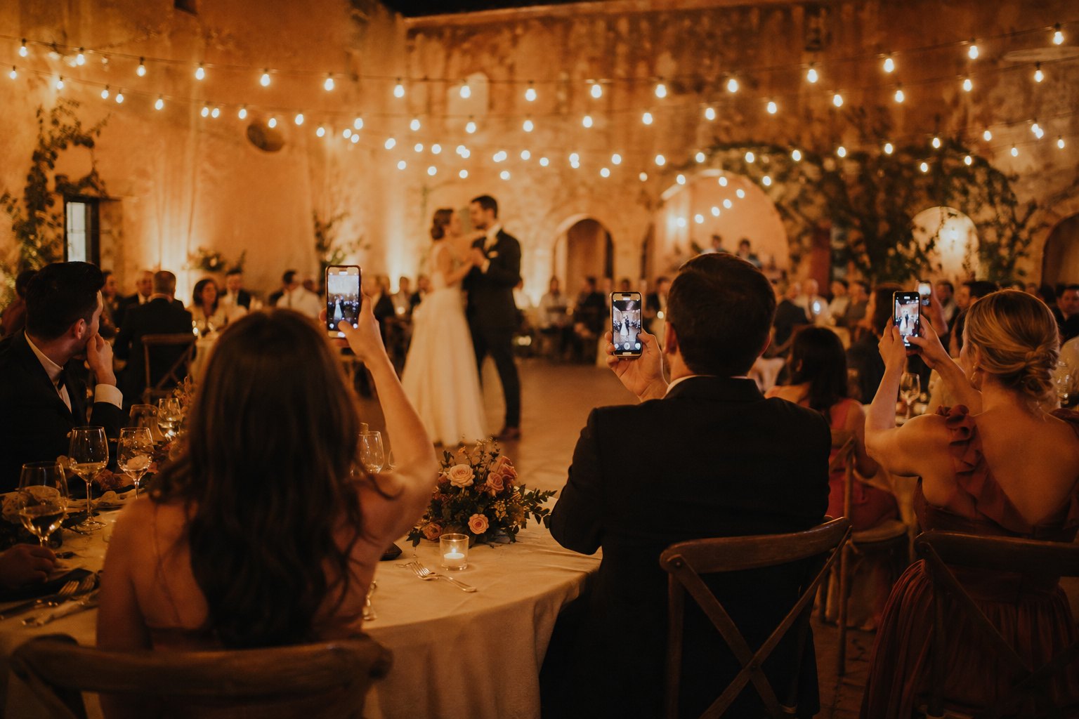 Wedding guests at a reception table holding their phones up to photograph the couple's first dance